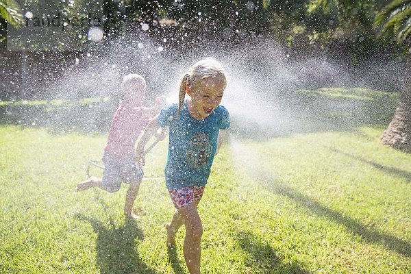 Junge läuft dem Mädchen im Garten hinterher  mit Wasserberieselung