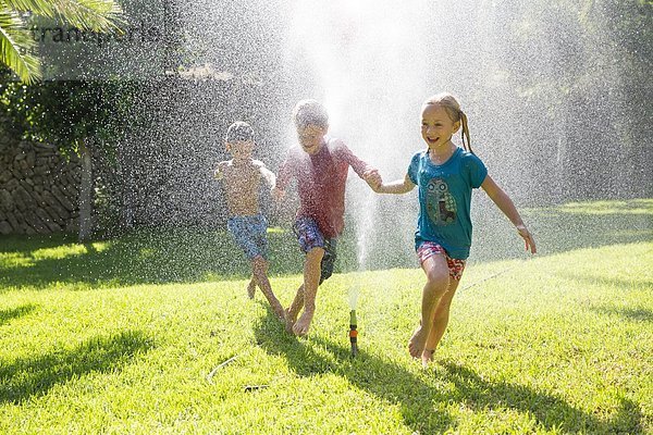 Drei Kinder im Garten  die durch eine Sprinkleranlage laufen.