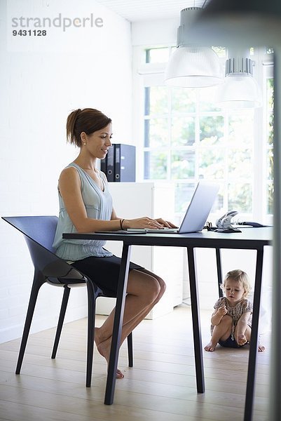 Mittlere erwachsene Mutter beim Tippen auf dem Laptop mit Kleinkind-Tochter unter dem Tisch