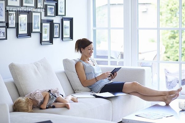 Mittlere erwachsene Frau mit digitalem Tablett auf dem Wohnzimmersofa  während die kleine Tochter schläft.