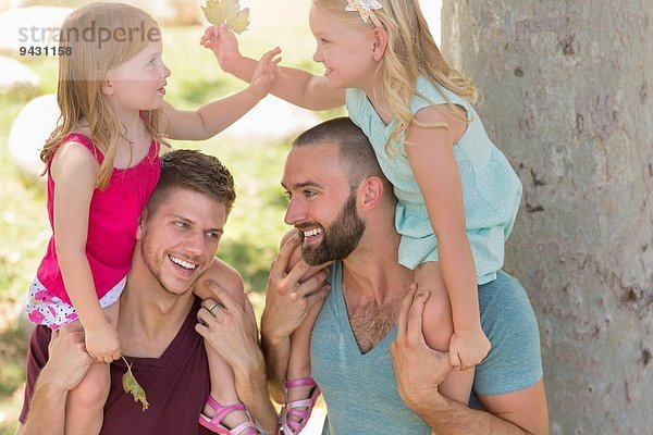 Zwei Schwestern sitzen auf den Schultern des Vaters im Park.