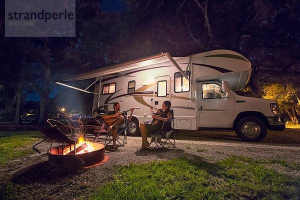 Mittleres erwachsenes Paar und kleine Tochter sitzen nachts vor dem Lagerfeuer.