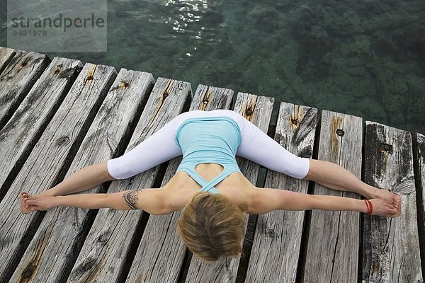 Overhead-Ansicht einer erwachsenen Frau mit ausgestreckten Armen und Beinen  die Yoga auf einer hölzernen Seebrücke praktiziert.