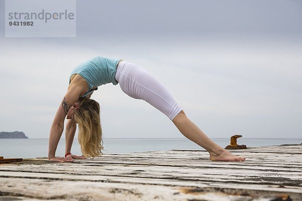 Mittlere erwachsene Frau  die sich nach hinten beugt und Yoga auf einem hölzernen Pier praktiziert.