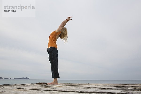 Mittlere erwachsene Frau  die sich zurückstreckt und Yoga auf einer hölzernen Seebrücke praktiziert.