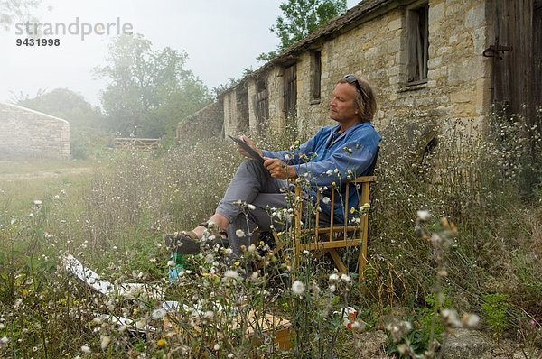 Mann sitzend skizzierend vor Bauernhofgebäuden