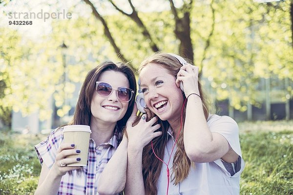 Zwei junge Freundinnen trinken Kaffee und hören Musik über Kopfhörer im Park.