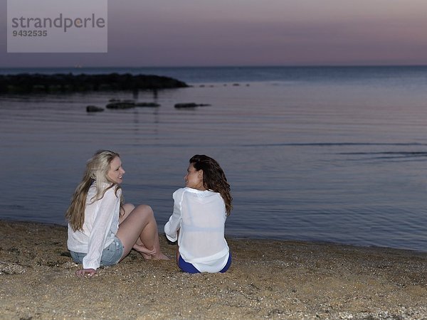 Rückansicht von zwei jungen Frauen  die bei Sonnenuntergang am Strand sitzen  Williamstown  Melbourne  Australien