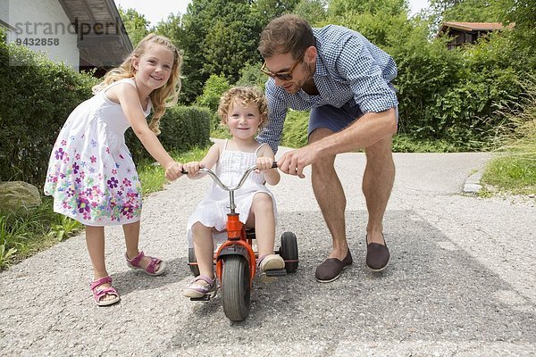 Mädchen auf dem Dreirad mit Vater und Schwester