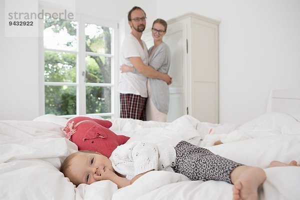 Kleines Mädchen auf dem Bett liegend  Eltern im Hintergrund