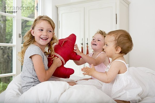 Drei Mädchen spielen auf dem Bett mit Plüschtier.