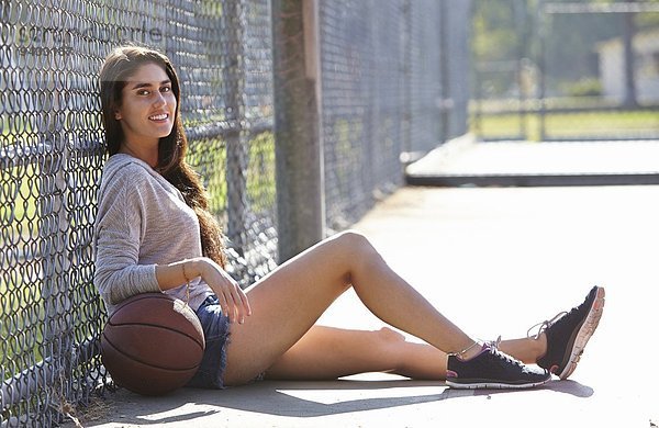 Junge Frau mit Basketball am Drahtzaun sitzend