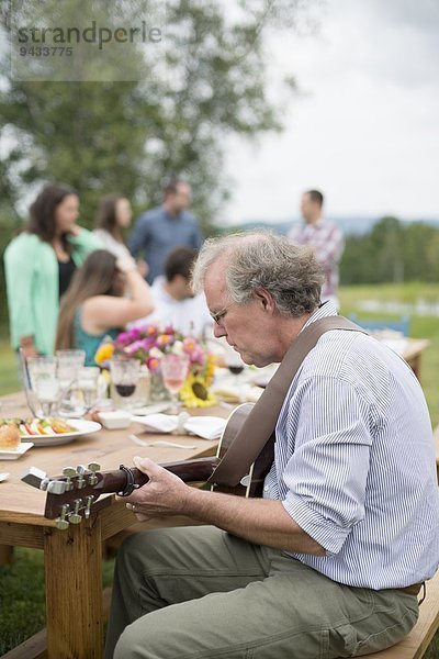Erwachsener Mann spielt Gitarre  während Freunde und Familie nach dem Essen miteinander reden  im Freien