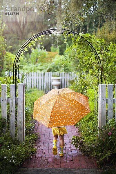 Mädchen geht durch das Gartentor und trägt einen Regenschirm.