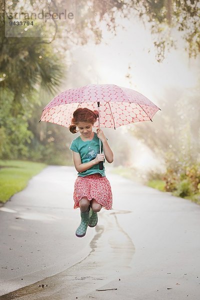 Mädchen hält Regenschirm hoch und springt Pfützen auf die Straße