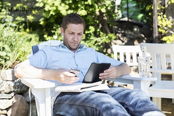 Mittlerer Erwachsener Mann mit digitalem Tablett beim Entspannen auf dem Liegestuhl im Hof