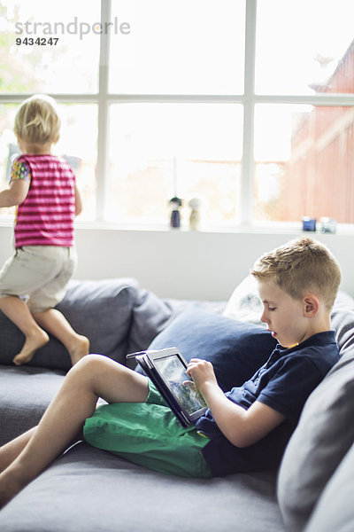 Junge mit digitalem Tablett auf dem Sofa  Schwester spielt im Hintergrund