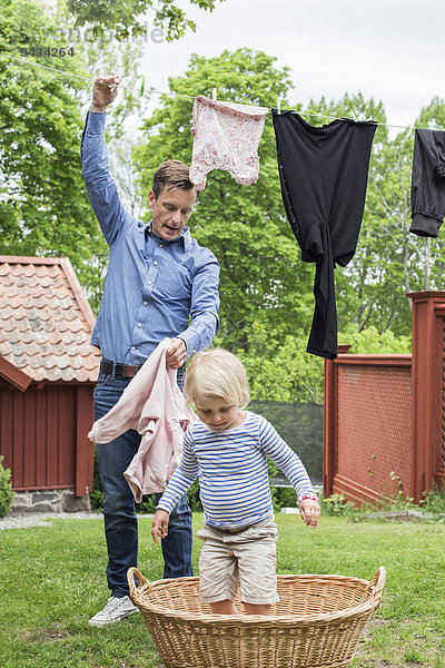 Mädchen steht im Wäschekorb  während Vater die Wäsche von der Wäscheleine nimmt.