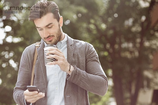 Junger Geschäftsmann beim Kaffeetrinken mit dem Smartphone im Freien