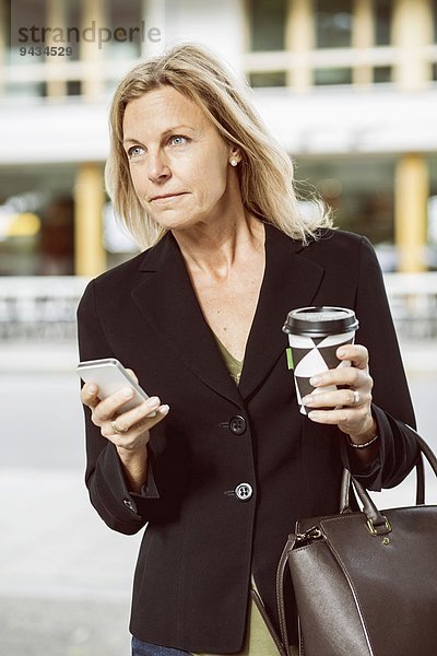 Geschäftsfrau mit Smartphone und Einweg-Kaffeetasse mit Blick nach draußen