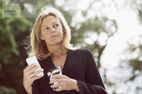 Geschäftsfrau schaut weg  während sie Smartphone und Einweg-Kaffeetasse im Freien hält.