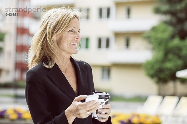 Glückliche Geschäftsfrau schaut weg  während sie Smartphone und Einweg-Kaffeetasse im Freien hält.