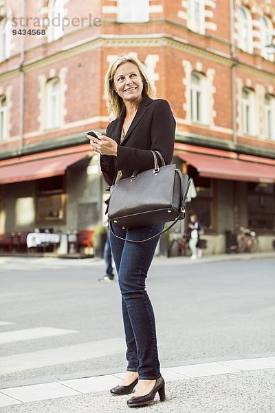 Volle Länge der lächelnden Geschäftsfrau  die weg schaut  während sie das Handy auf der Straße benutzt.