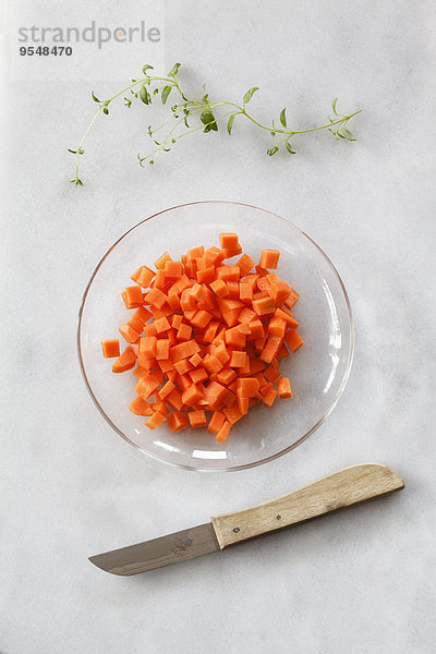 Glasschale mit Karottenwürfeln und Küchenmesser auf weißem Grund