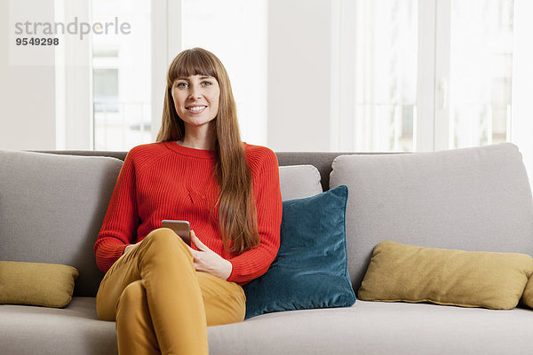 Lächelnde Frau sitzt auf dem Sofa und hält das Handy.