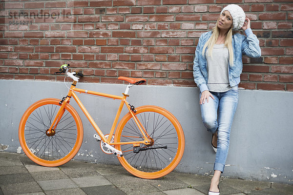 Modische junge Frau lehnt sich an die Ziegelwand neben dem orangefarbenen Fahrrad.