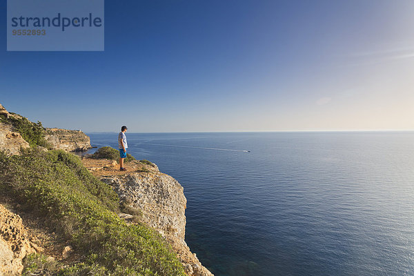 Spanien  Balearen  Mallorca  ein Teenager  der auf einem Felsen an der Steilküste steht und den Sonnenuntergang beobachtet.