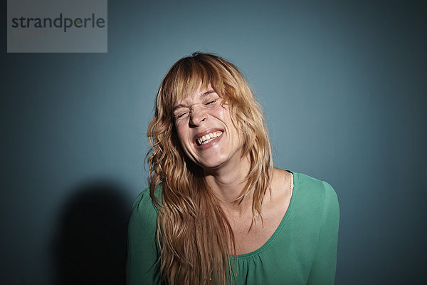 Porträt einer lachenden blonden Frau mit geschlossenen Augen vor blauem Hintergrund