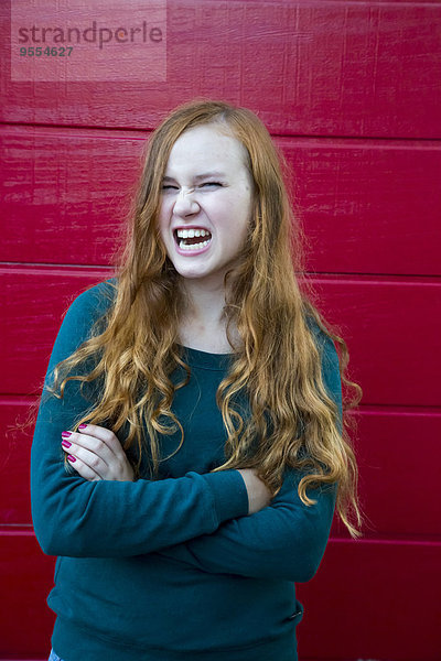 Porträt eines wütenden Teenagermädchens mit gekreuzten Armen vor roter Holzwand