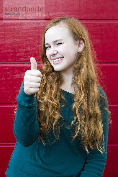 Porträt eines lächelnden Teenagermädchens mit Daumen vor roter Holzwand