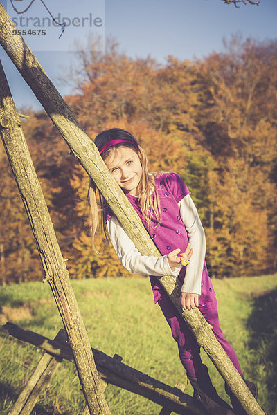 Porträt eines lächelnden kleinen Mädchens  das sich vor dem Herbstwald auf eine Holzstange stützt.