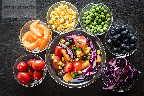 Gemischte Salat- und Glasschalen mit verschiedenen Rohkost-Regenbogenfarben auf Schiefer angeordnet