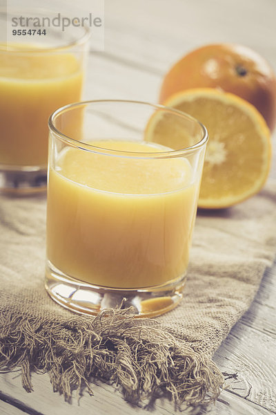 Zwei Gläser frisch gepresster Orangensaft und Orangen auf Tuch und Holz