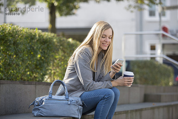 Lächelnde junge Frauen sitzen auf Stufen mit Kaffee zum Mitnehmen mit dem Smartphone