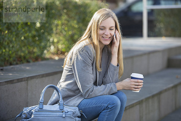 Lächelnde junge Frauen sitzen auf der Treppe mit Kaffee zum Telefonieren mit dem Smartphone.