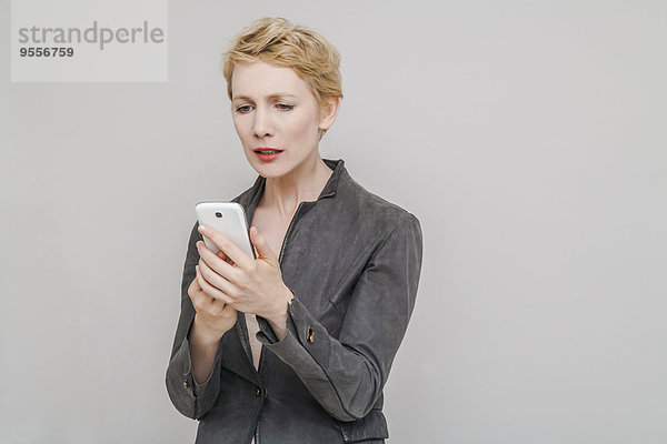 Porträt einer verstörten blonden Frau  die ihr Smartphone vor grauem Hintergrund anschaut