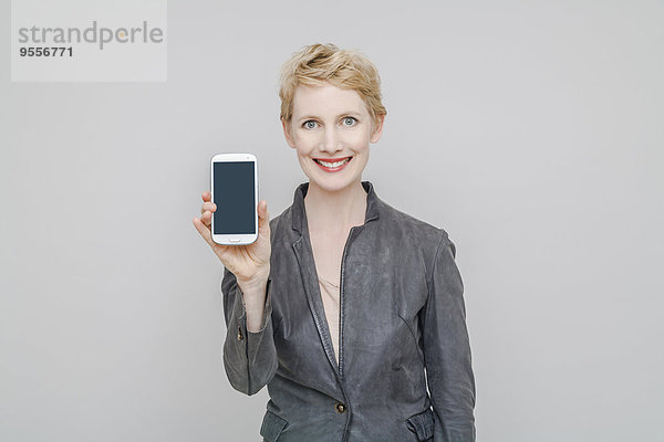 Porträt einer lächelnden blonden Frau  die ihr Smartphone vor grauem Hintergrund zeigt.