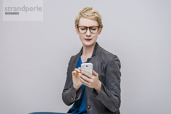 Porträt einer lächelnden blonden Frau mit Smartphone vor grauem Hintergrund