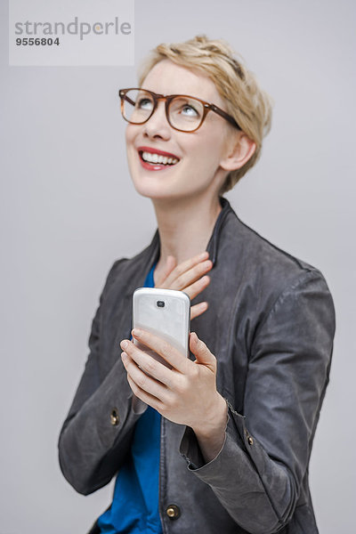 Porträt einer lächelnden blonden Frau mit ihrem Smartphone vor grauem Hintergrund