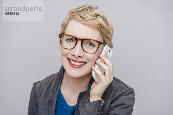 Porträt einer lächelnden  blonden Frau mit Brille  die vor grauem Hintergrund telefoniert.