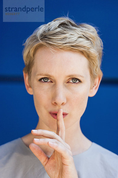 Porträt einer blonden Frau mit Finger auf dem Mund vor blauem Hintergrund