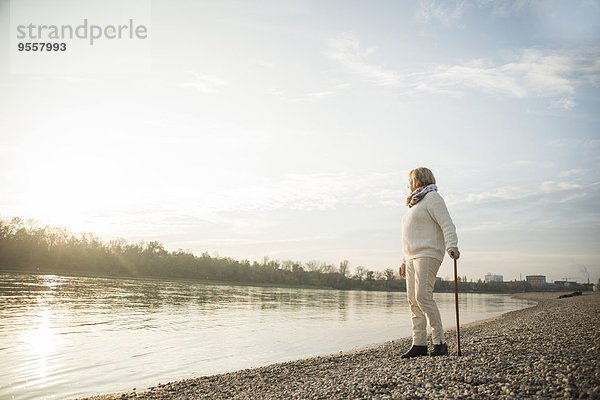 Seniorenfrau mit Spazierstock am Wasser stehend  Sonnenuntergang beobachtend