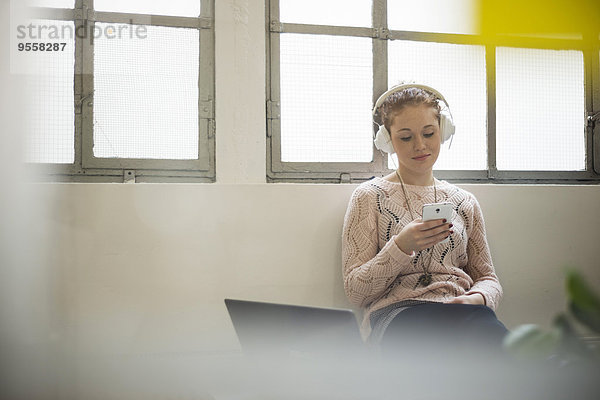Junge Frau auf dem Schreibtisch sitzend mit Kopfhörer und Handy