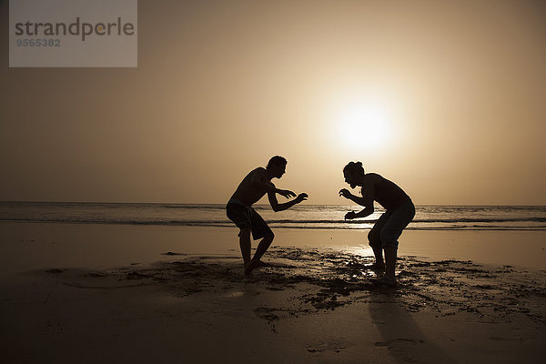 Volle Länge der Silhouette männliche Freunde spielen am Strand bei Sonnenuntergang
