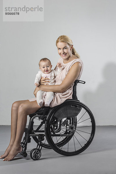 Portrait einer glücklichen Frau mit einem Mädchen im Rollstuhl vor grauem Hintergrund.