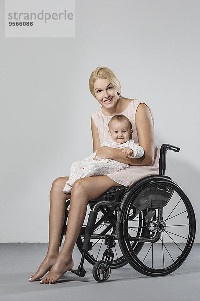 Ganzflächiges Porträt einer glücklichen Frau mit Tochter im Rollstuhl vor grauem Hintergrund